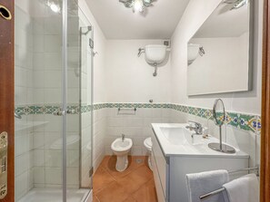 Baño con ducha de vidrio, lavamanos pedestal, inodoro y espejo sobre el lavamanos.