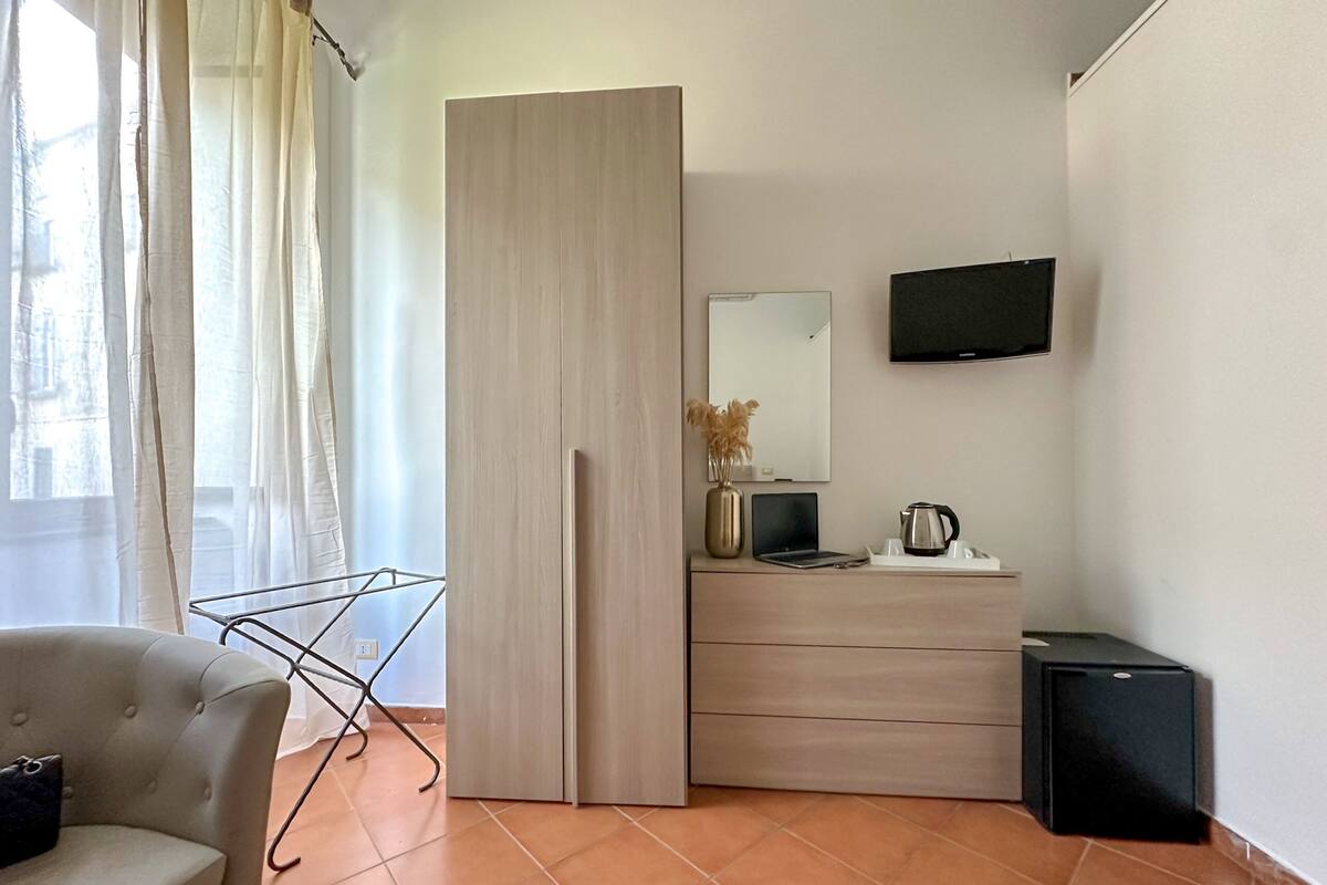A modern hotel room with a sofa, a wardrobe, a flat-screen TV, a small table with a vase and a kettle, and a compact refrigerator.