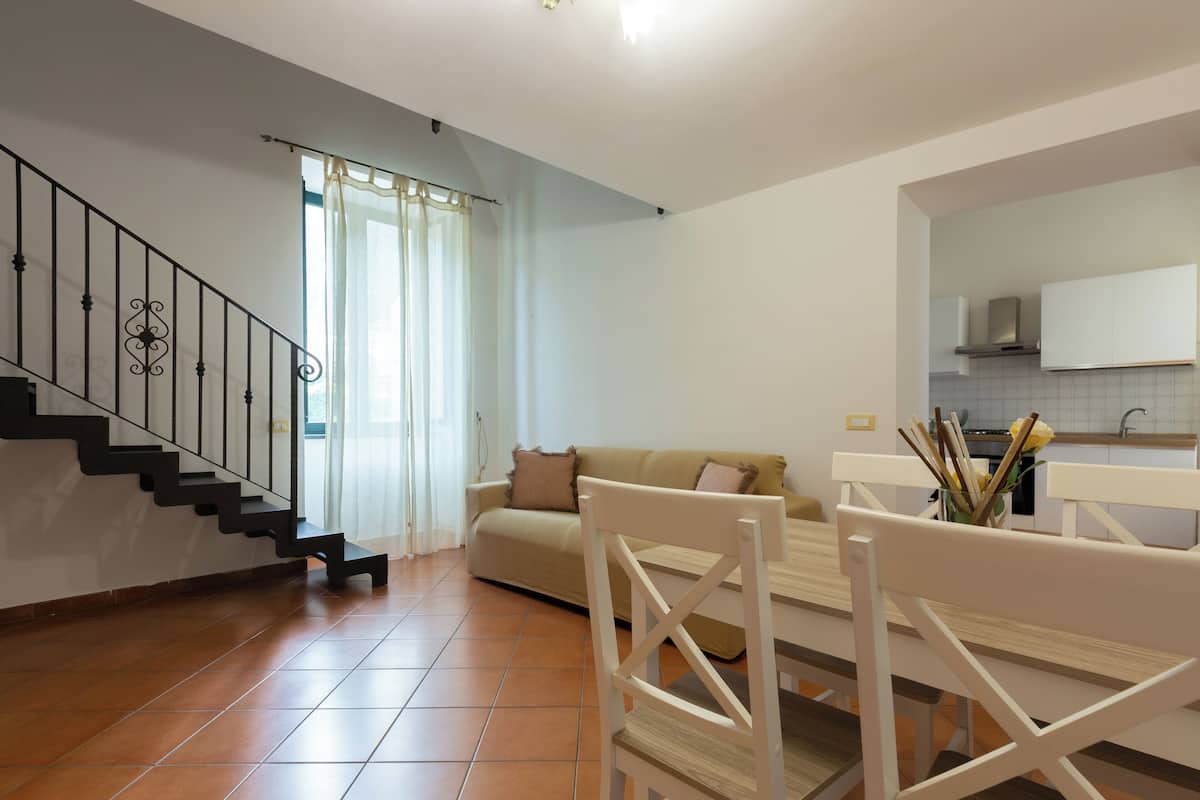 A spacious living area with a staircase, a sofa, a dining table with chairs, and a kitchen in the background.