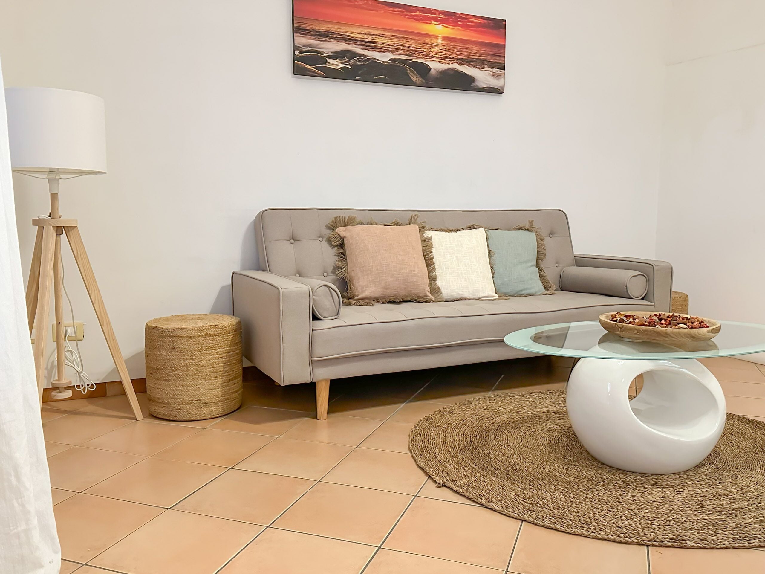 A modern living room with a grey sofa, a glass coffee table, a woven rug, a lamp, and a painting of a sunset.