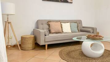 A modern living room with a grey sofa, a glass coffee table, a woven rug, a lamp, and a painting of a sunset.