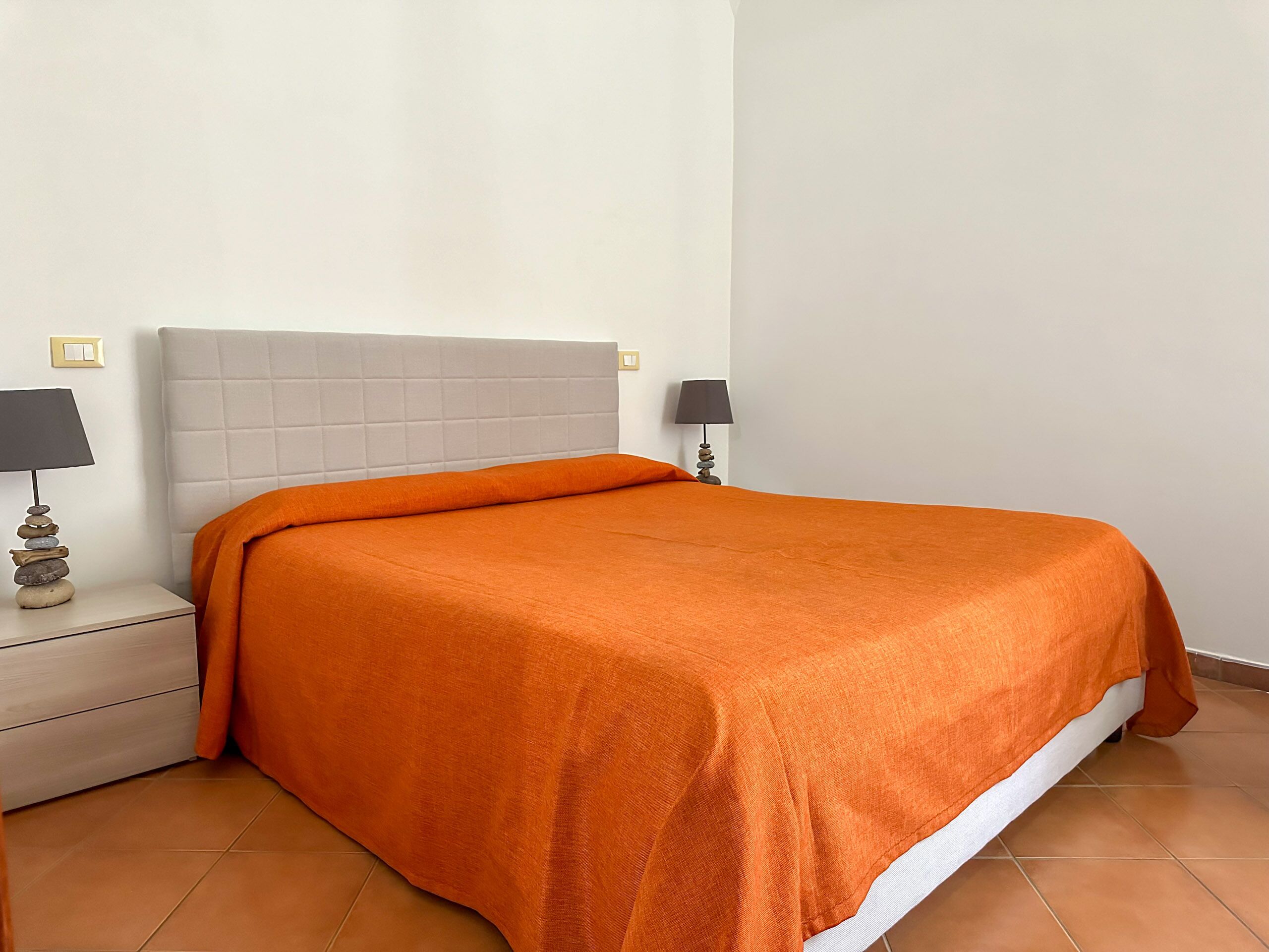 Ein ordentlich bezogenes Bett mit einer orangefarbenen Decke, einem weißen Kopfteil und einem Nachttisch mit Lampe.