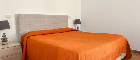 Una cama bien hecha con una manta naranja, un cabecero blanco y una mesita de noche con una lámpara.