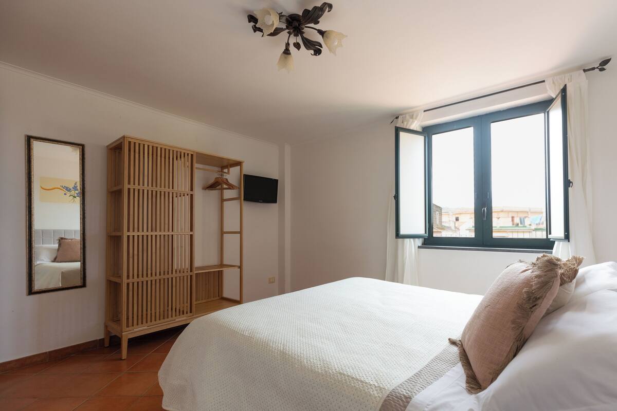A bedroom with a bed, a wooden wardrobe, a television, and a window with curtains.
