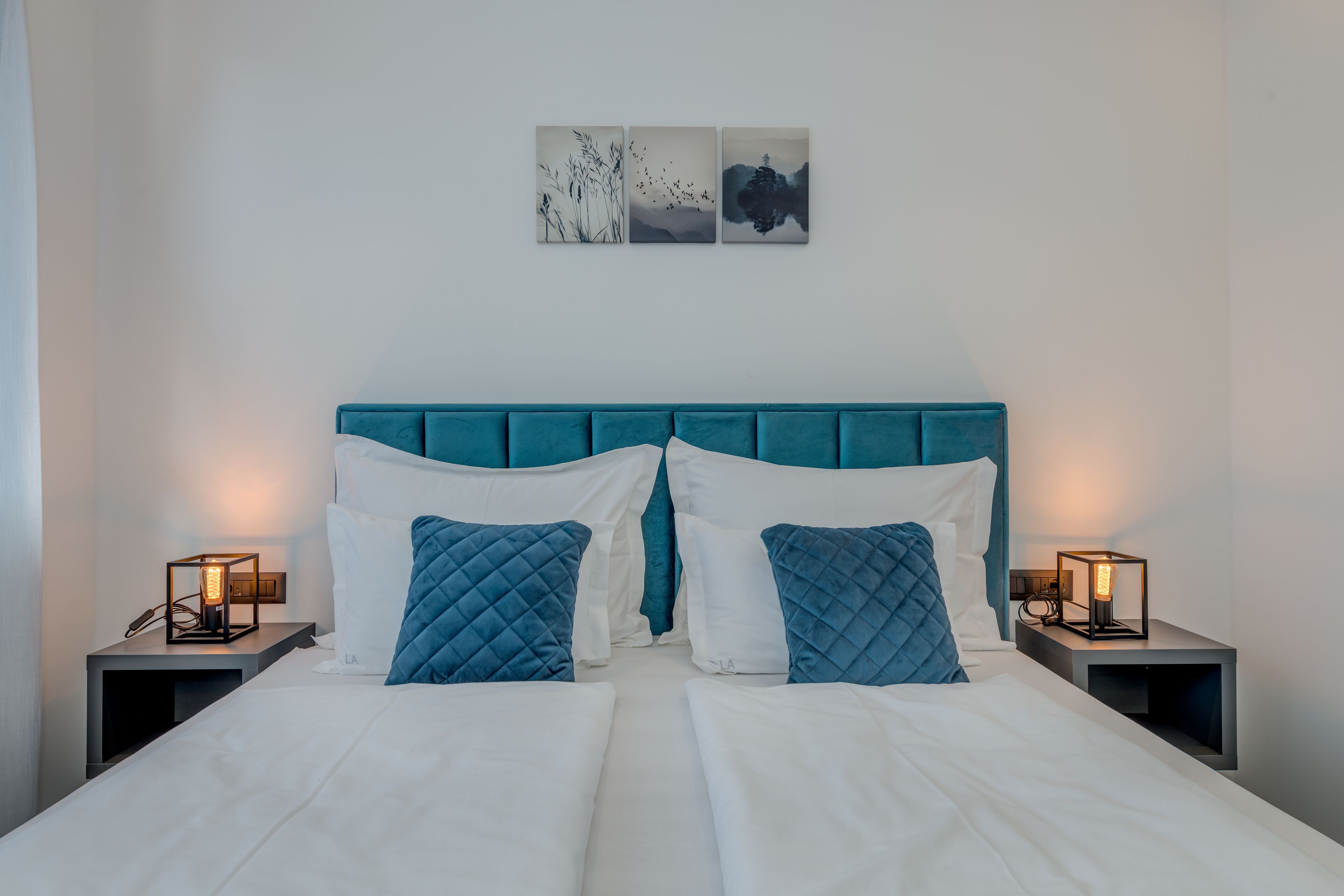 A double bed with a teal headboard, two bedside tables with lamps, and three wall art pieces above the bed.