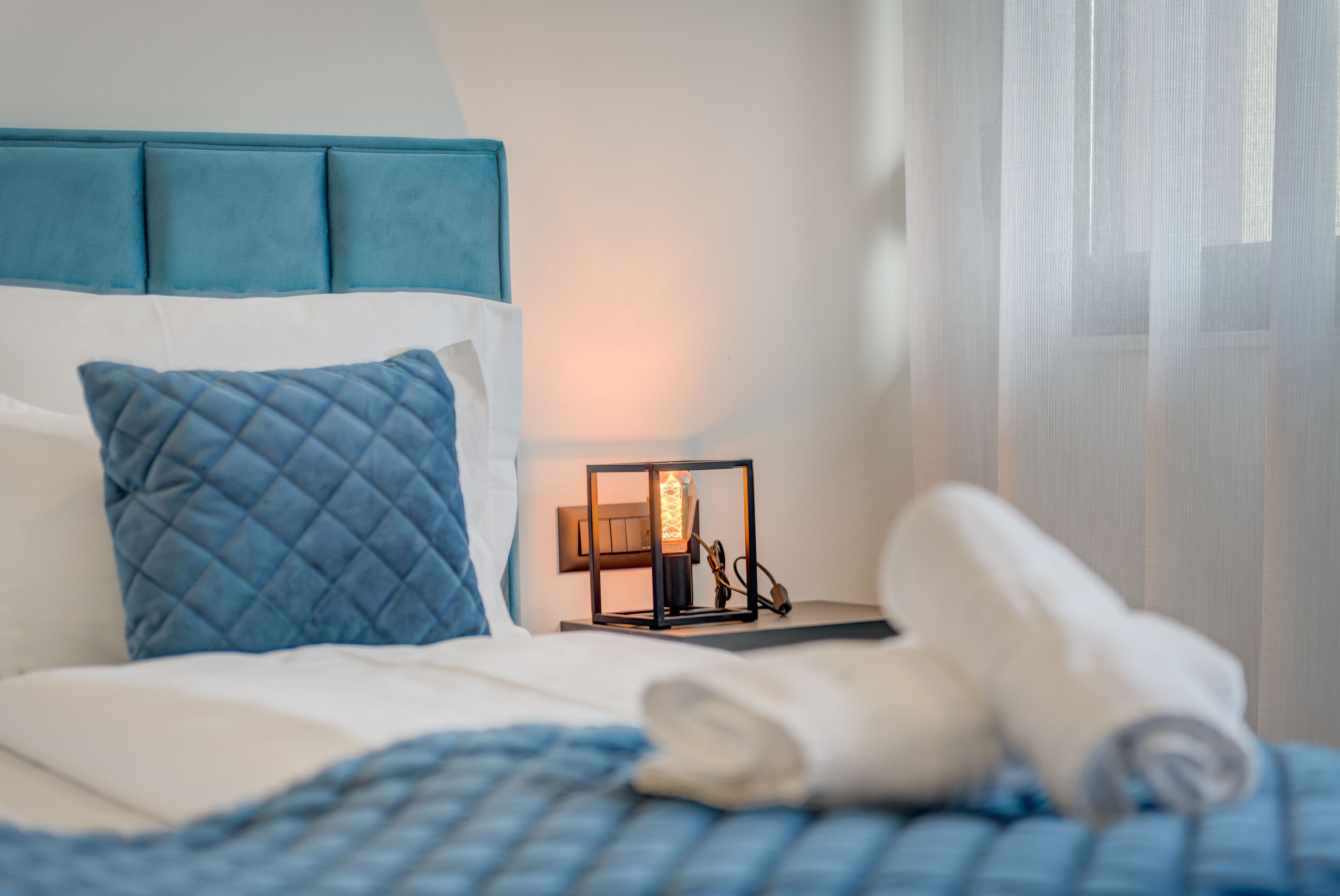Un lit bien fait, avec un oreiller matelassé, une lampe de chevet et une serviette enroulée posée sur un couvre-lit blanc.