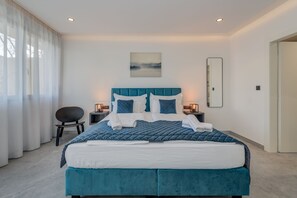 A modern bedroom with a large bed, a chair, bedside tables, and a painting on the wall.