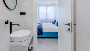 A modern bathroom with a white sink, a mirror, and a door leading to a bedroom with a bed and a window.