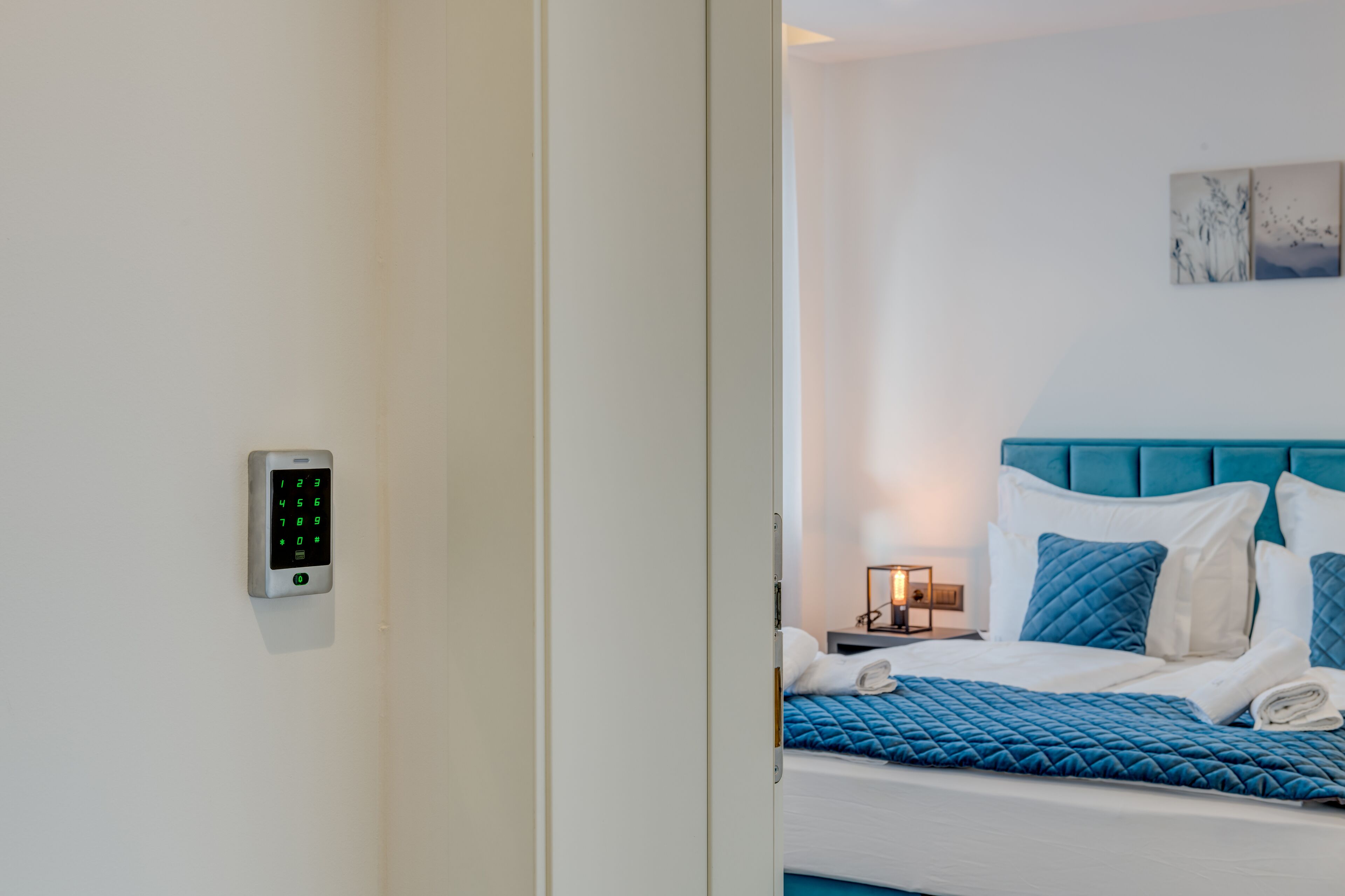 A hotel room with a bed, a bedside table, a wall-mounted control panel, and a nightstand with a lamp.