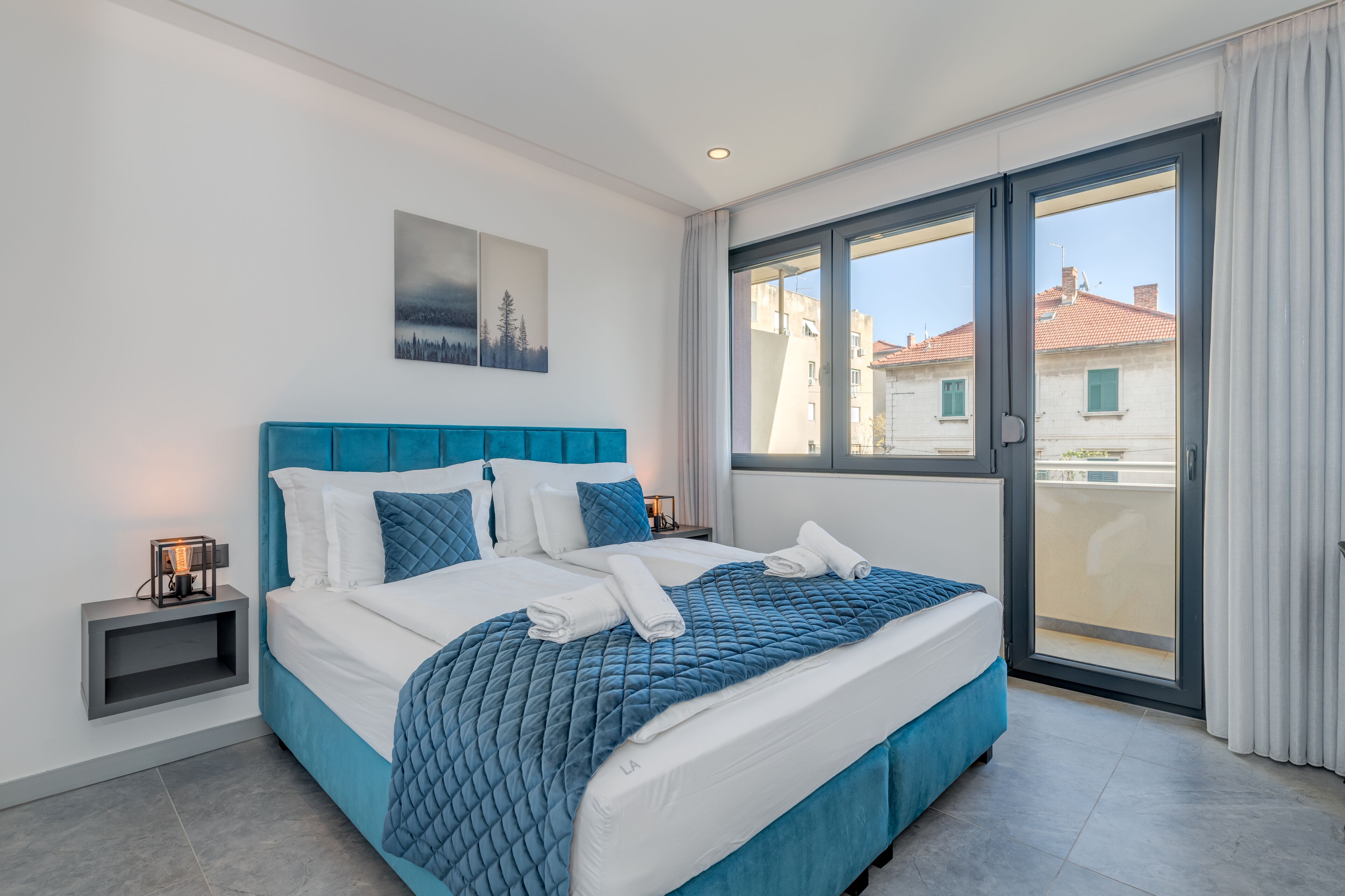 Ein modernes Schlafzimmer mit einem großen Bett, einem Nachttisch, einem Gemälde und Blick auf Gebäude durch ein großes Fenster.