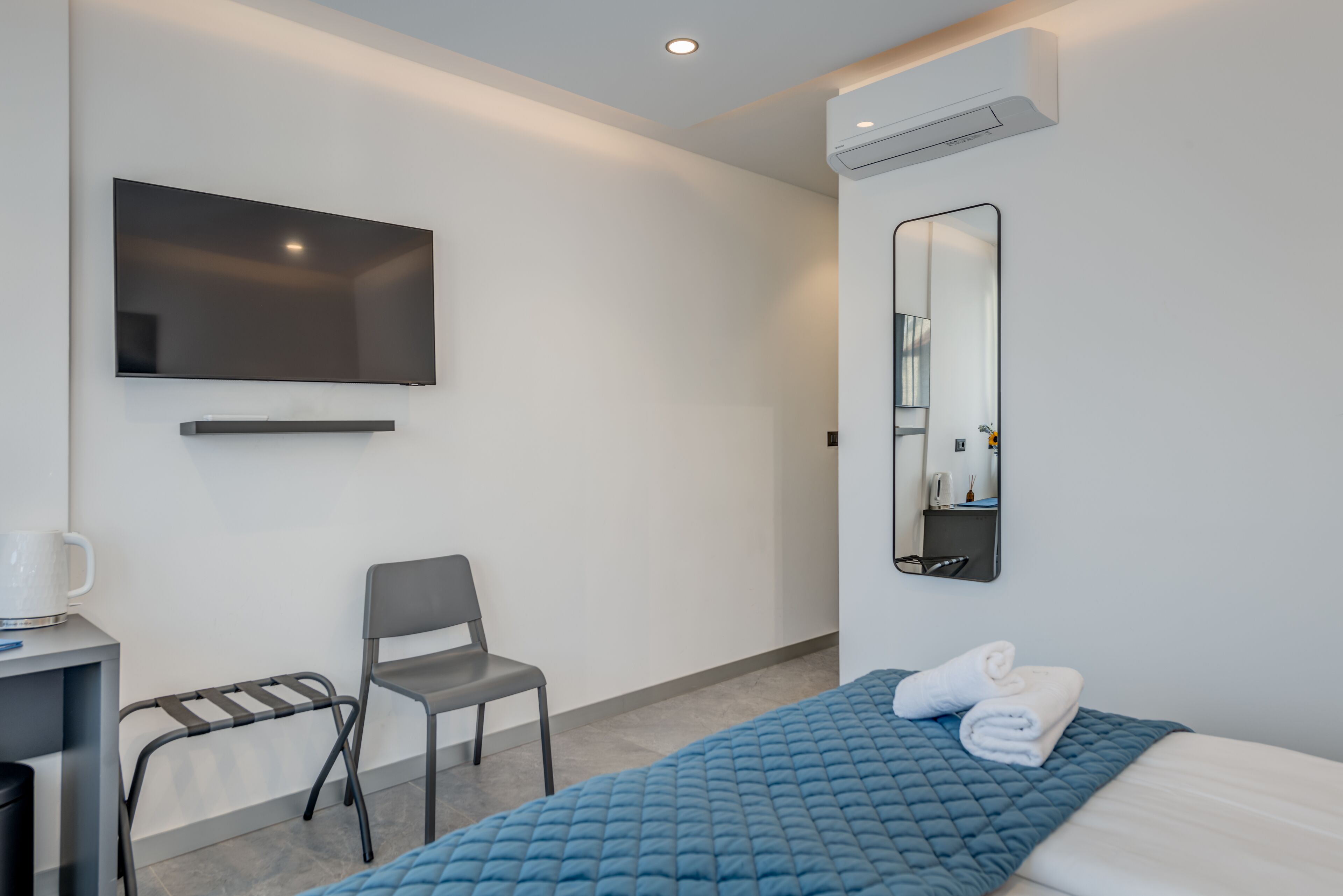 Habitación de hotel con cama, una silla, un escritorio con hervidor, un televisor de pantalla plana y un espejo.