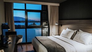 A hotel room with a large bed, a desk with a chair, a desk lamp, and a view of a body of water and mountains through a large window.