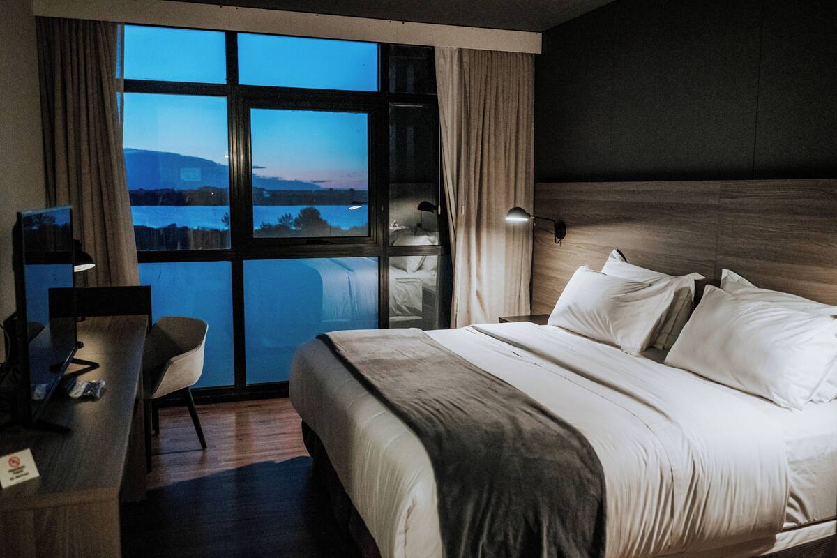 Une chambre d’hôtel avec un grand lit, un bureau avec une chaise, une lampe de bureau et une vue sur un plan d’eau et des montagnes, le tout visible à travers une grande fenêtre.