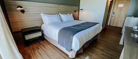 A modern hotel room with a large bed, wooden flooring, a desk, and a chair.