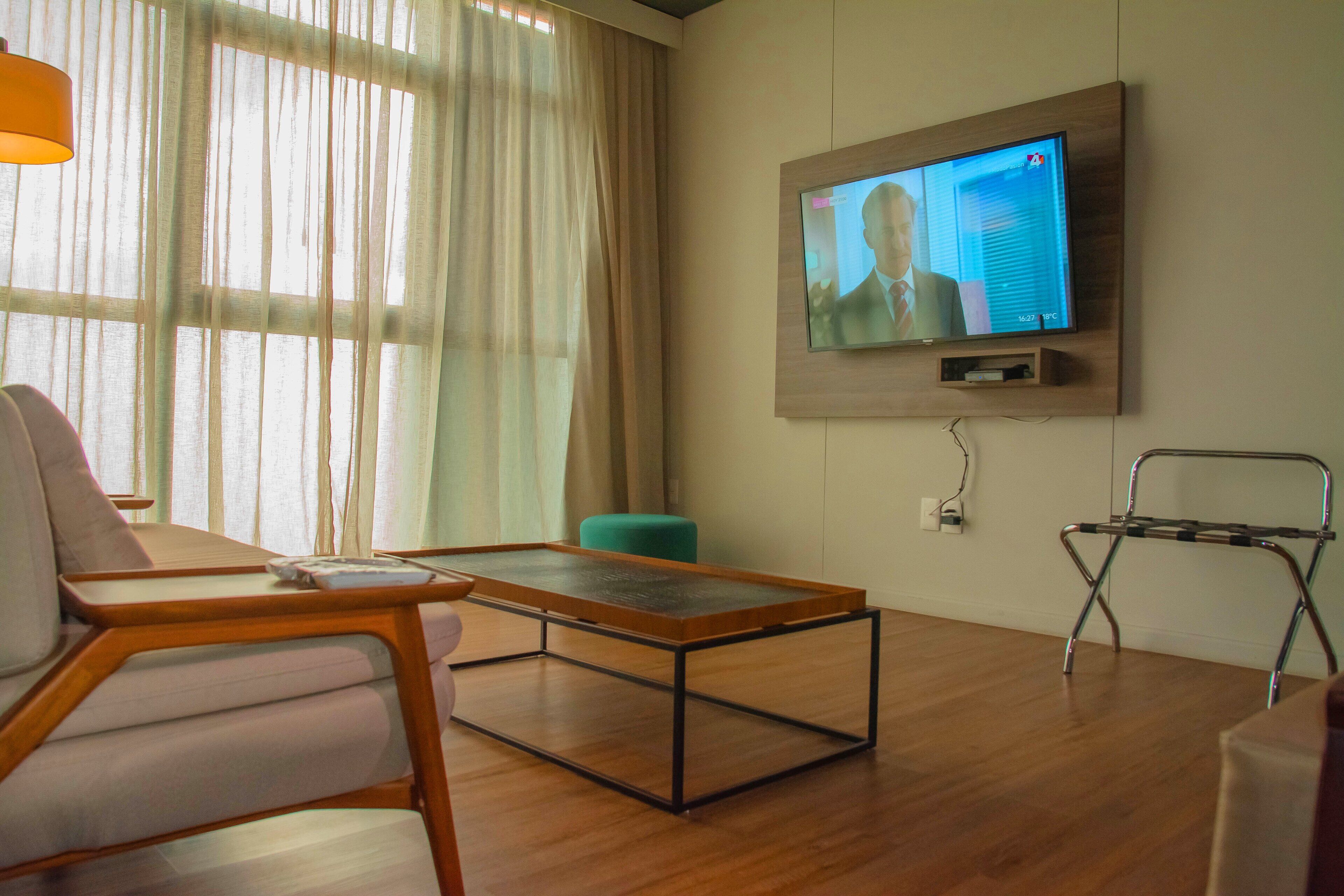 A modern living room with a wooden coffee table, a sofa, a teal ottoman, and a television mounted on the wall.