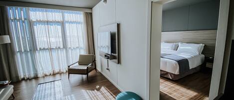 Quarto de hotel moderno com cama, televisão fixada na parede, área de estar com poltrona e uma pequena mesa.