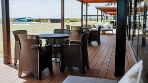 An outdoor patio with tables and chairs, a wooden deck, and a view of the beach and ocean.