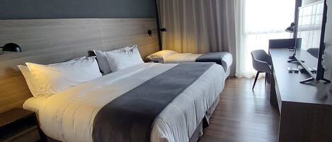 A modern hotel room with a large bed, a desk with a computer, and a window with curtains.