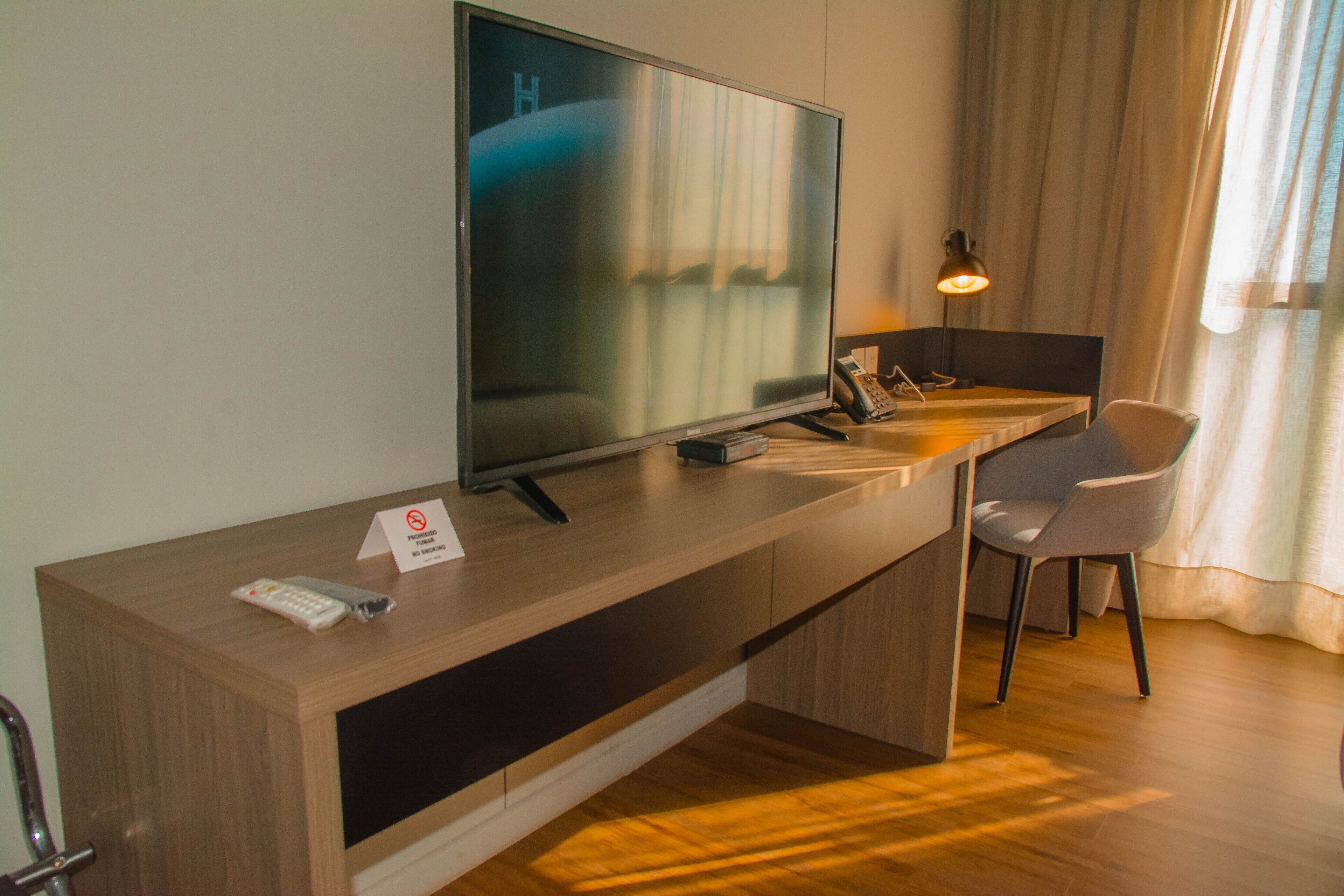 A hotel room with a wooden desk, a television, a chair, and a bedside table.