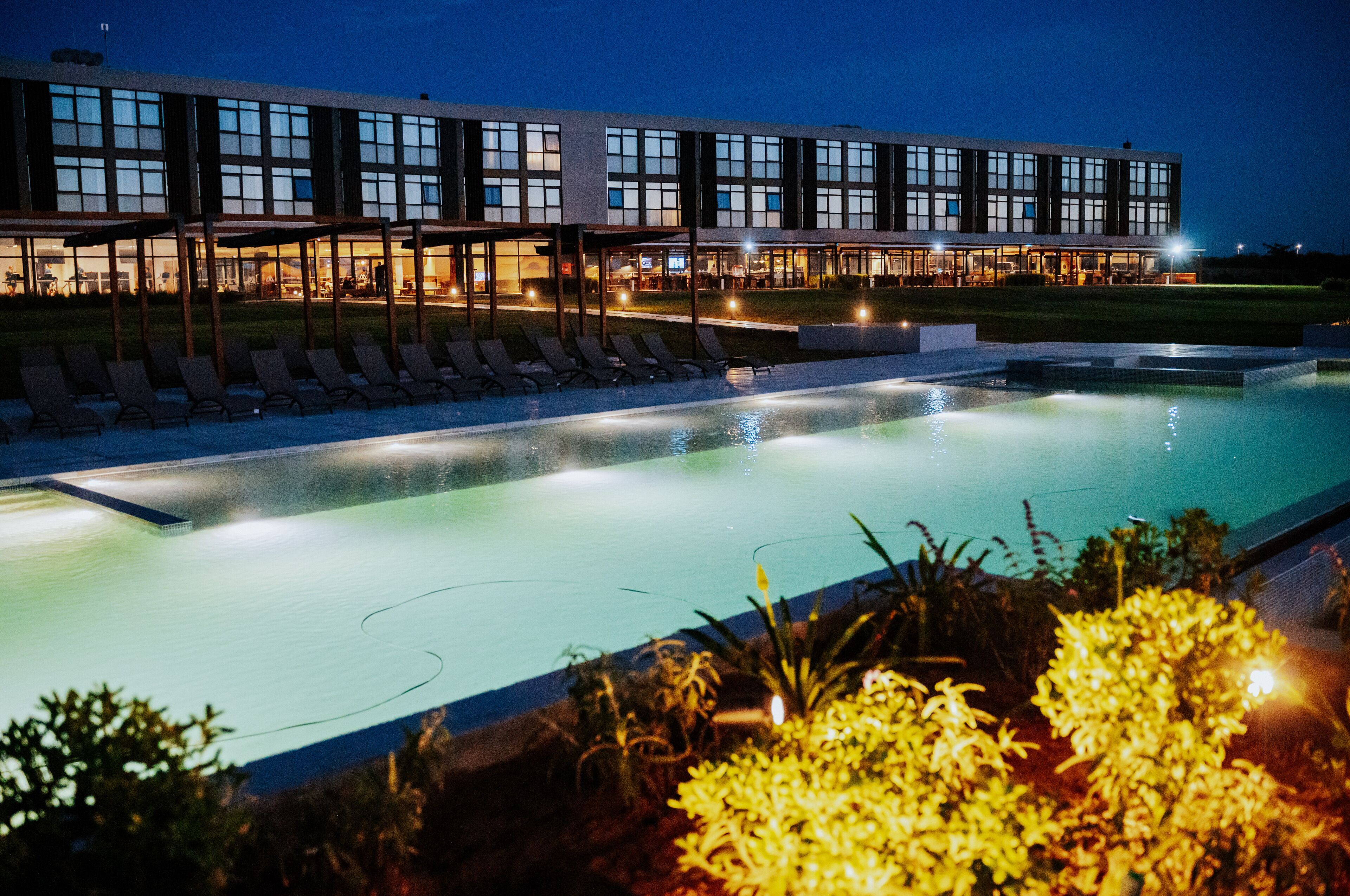 A modern hotel with a swimming pool, lounge chairs, and a well-lit building facade.
