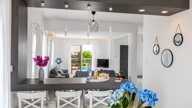 Una cocina moderna con comedor, sillas blancas, un florero y un televisor al fondo.
