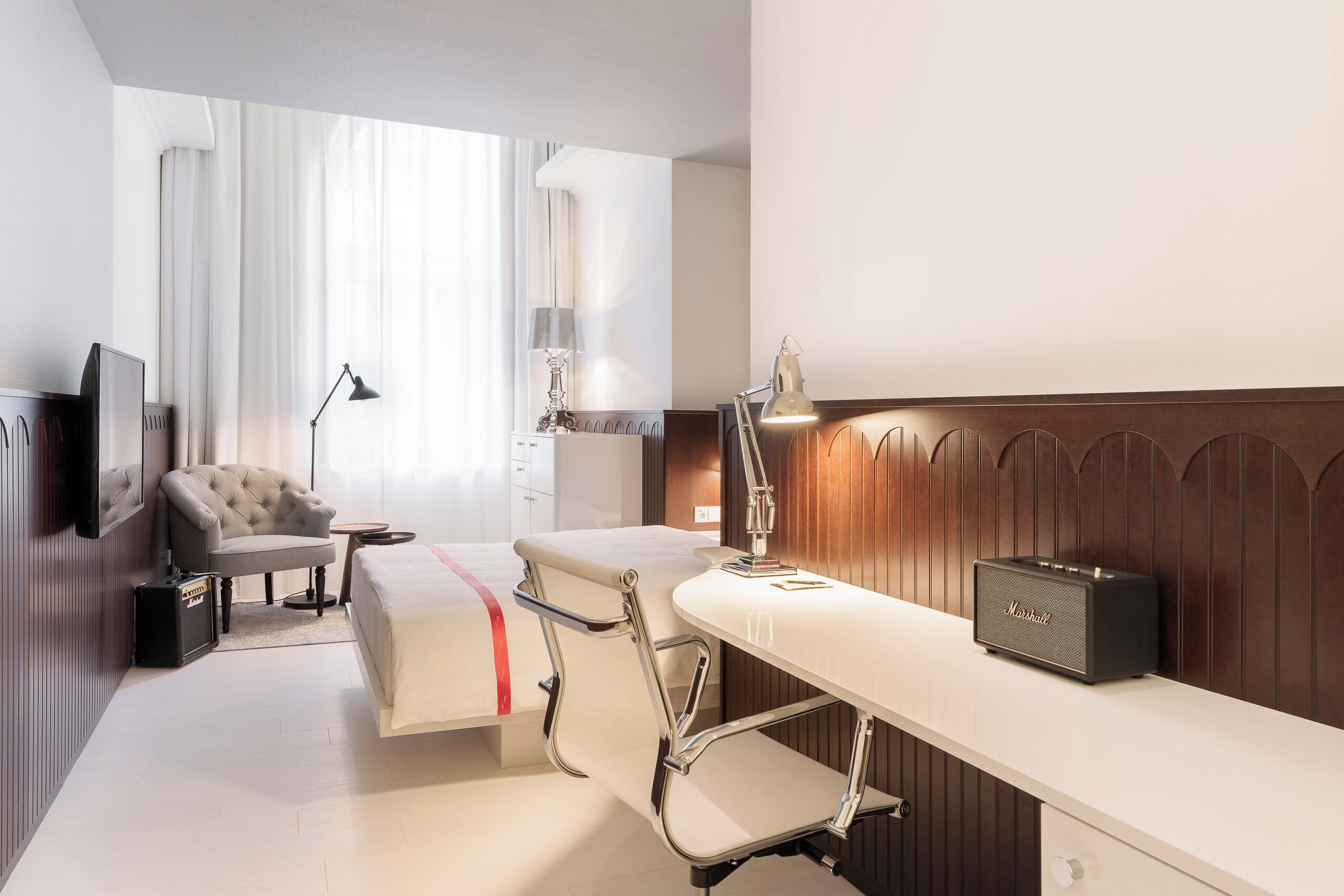 A hotel room with a bed, a desk, a chair, a lamp, a radio, and a suitcase.