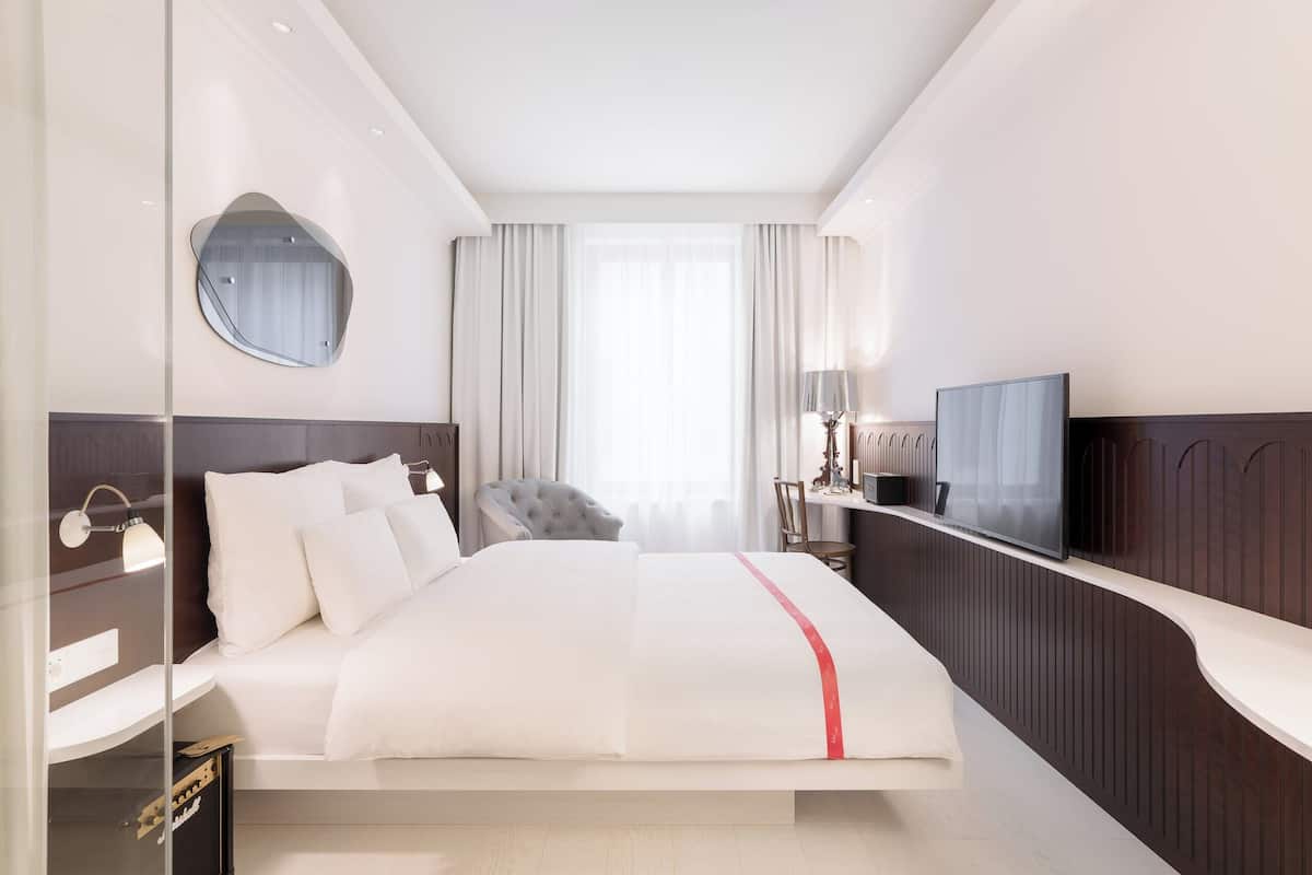 Una habitación de hotel moderna con una cama grande, televisión, espejo y una zona de estar con una silla.