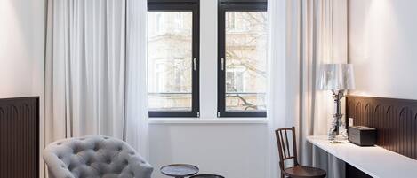 Una habitación con un sillón capitonado, dos pequeñas mesas redondas, una silla de madera, una lámpara y un radiador.