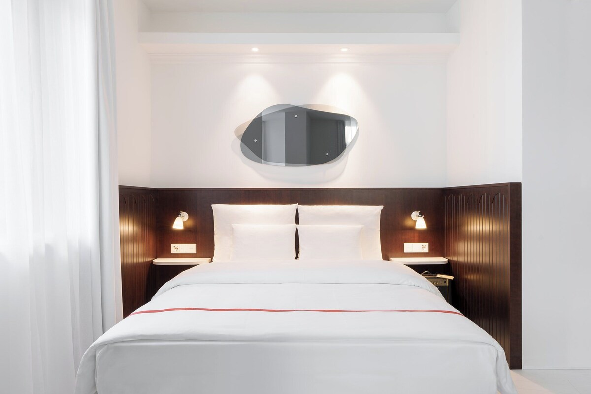 Habitación de hotel con una cama grande, dos mesitas de noche, un espejo sobre la cama y una pequeña mesita auxiliar.