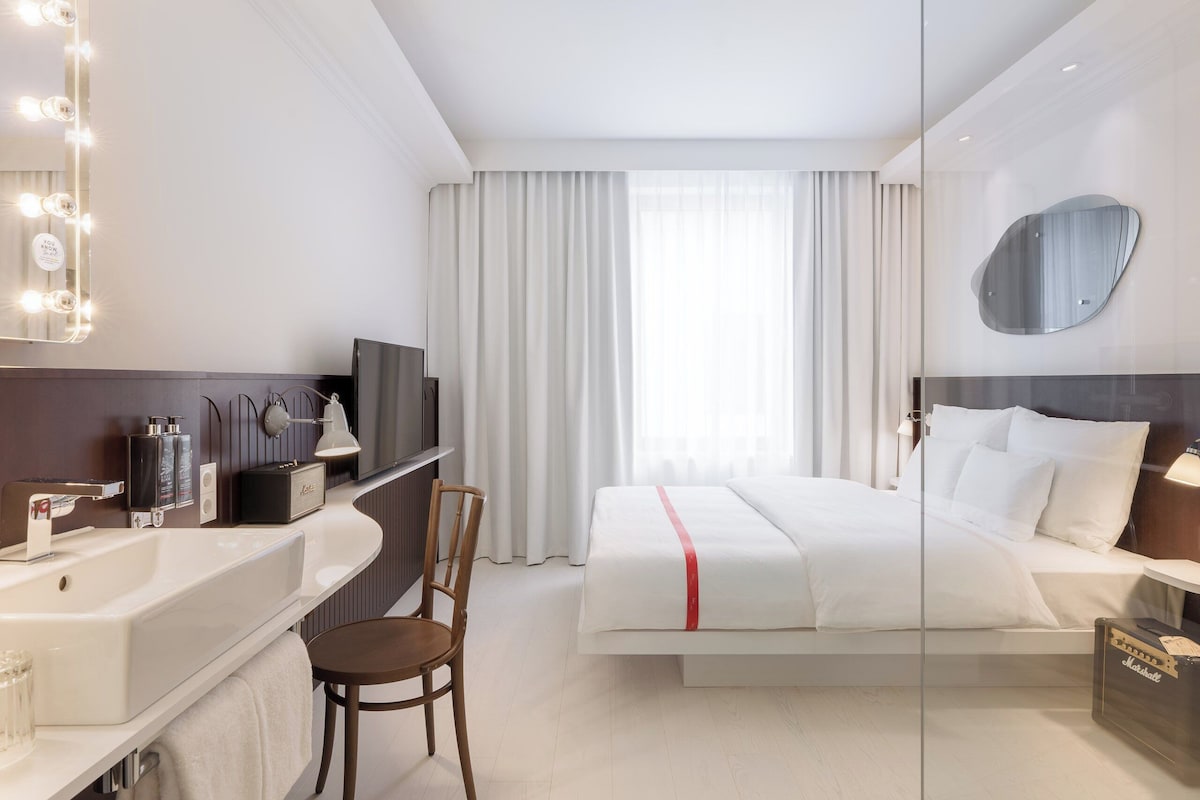A hotel room with a large bed, a vanity with a mirror and lights, a television, and a glass partition.