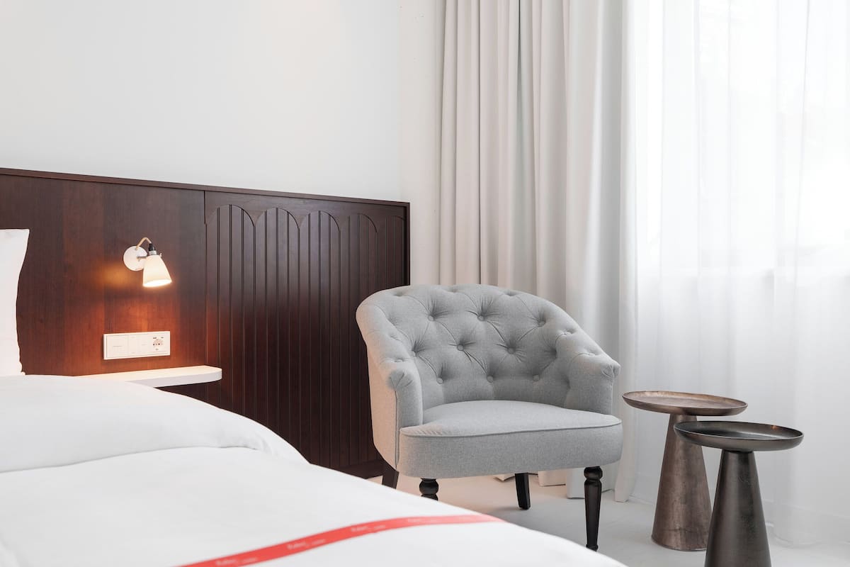 Habitación de hotel con cama, un sillón capitonado, dos mesitas de noche, una lámpara de pared y un ventanal con cortinas.