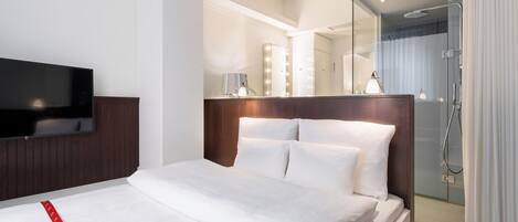 Habitación de hotel con cama, televisor, espejo con luces y zona de ducha.