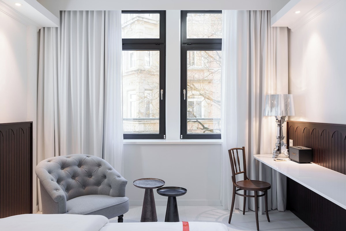 A hotel room with a bed, a grey armchair, a wooden chair, a small round table, a lamp, and a view of buildings through the window.