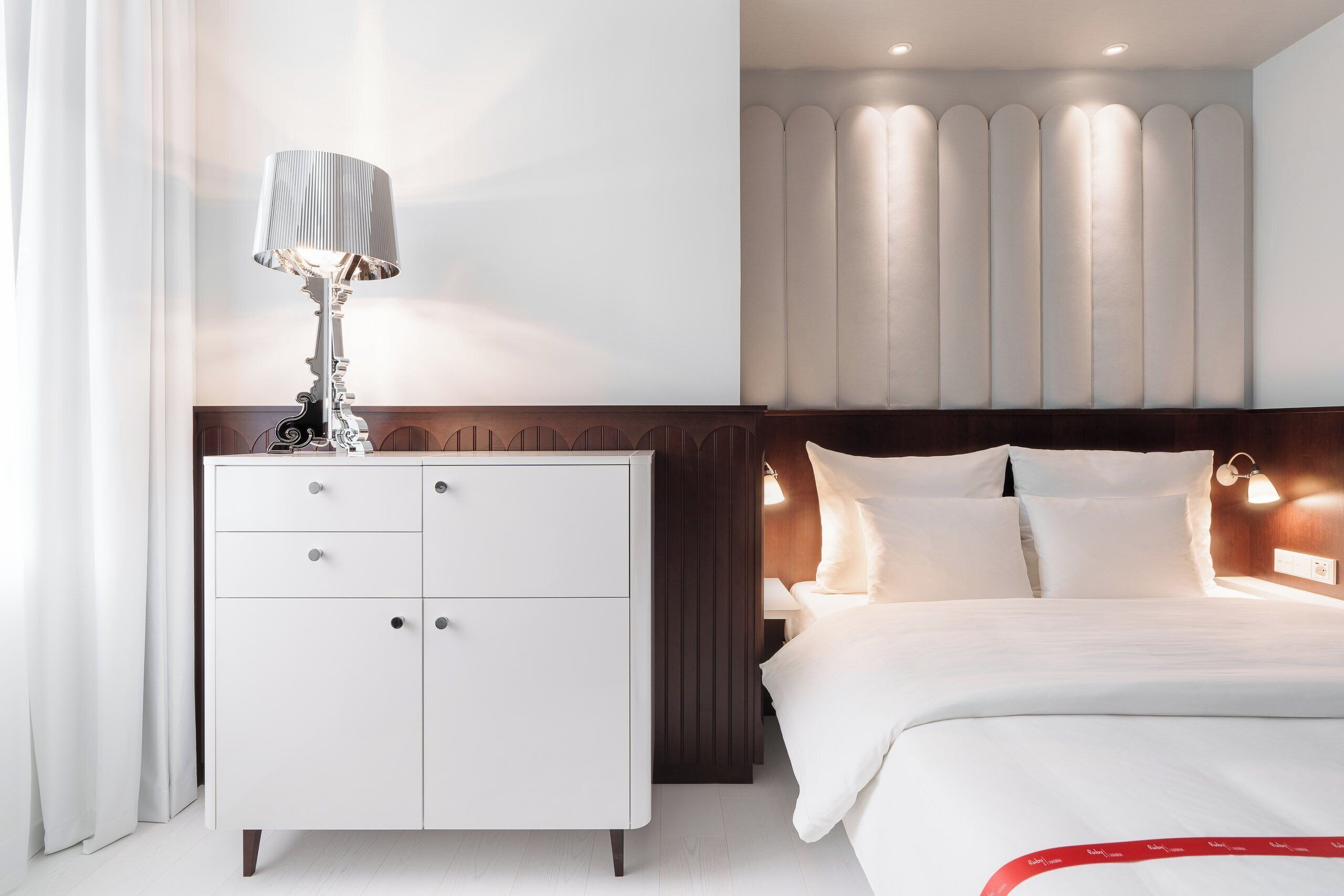 Una camera d'albergo con un letto, un comodino, una lampada e un punto luce a parete.