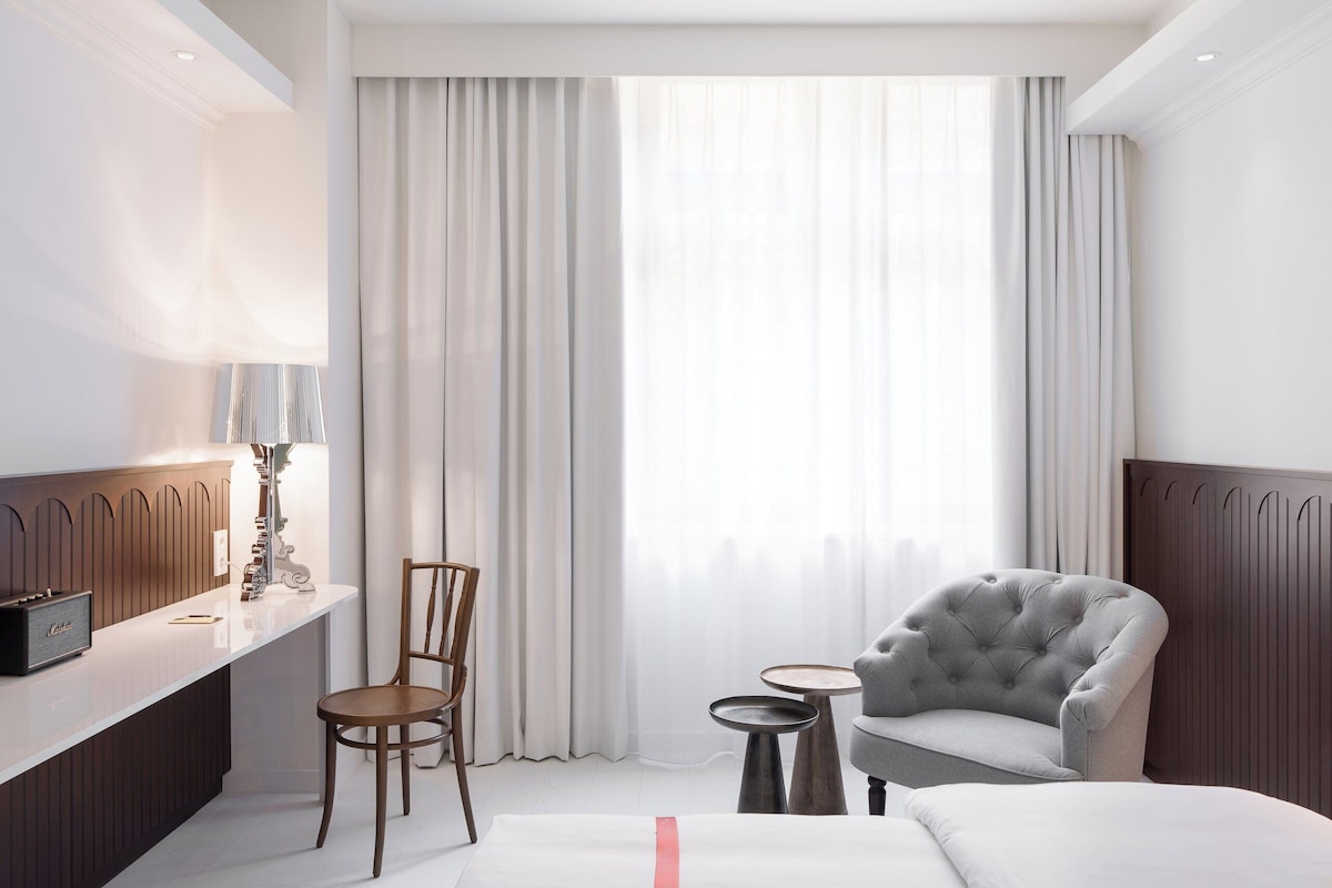 A hotel room with a bed, a chair, a small table, a lamp, and a window with curtains.