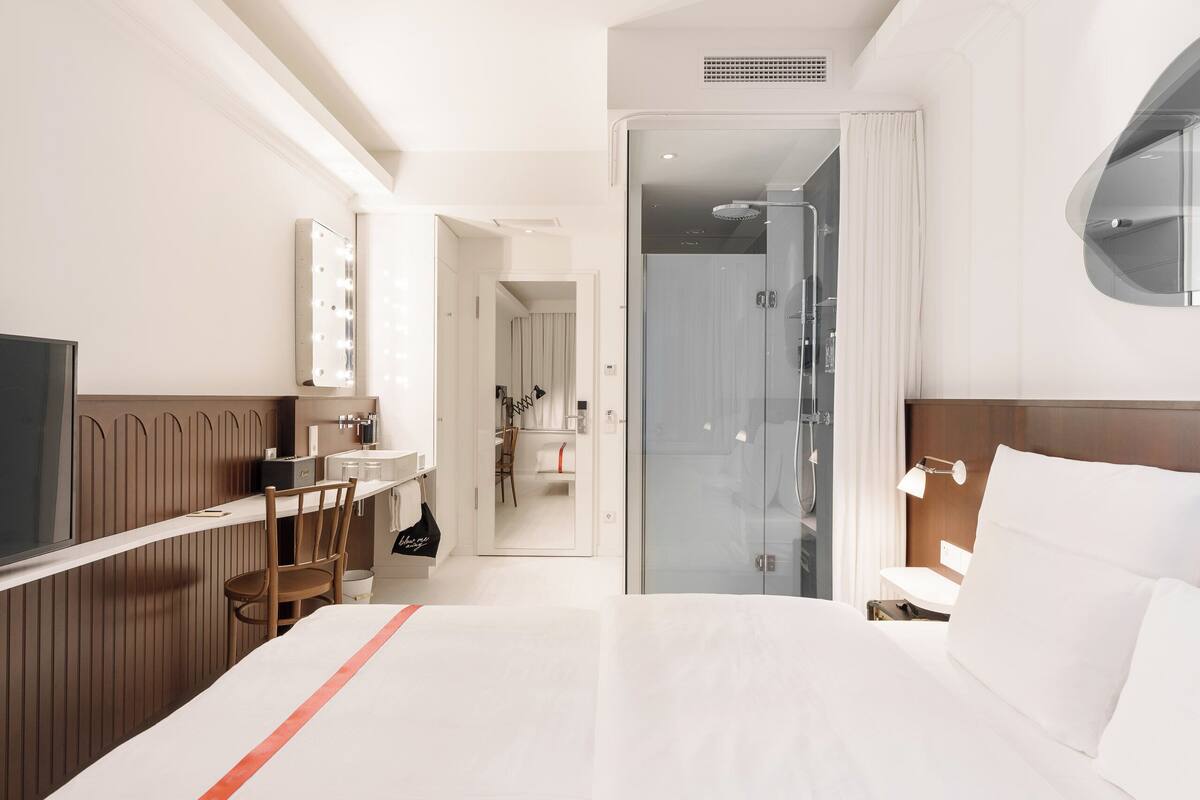 Habitación de hotel con cama, escritorio, silla, espejo, ducha y televisión.