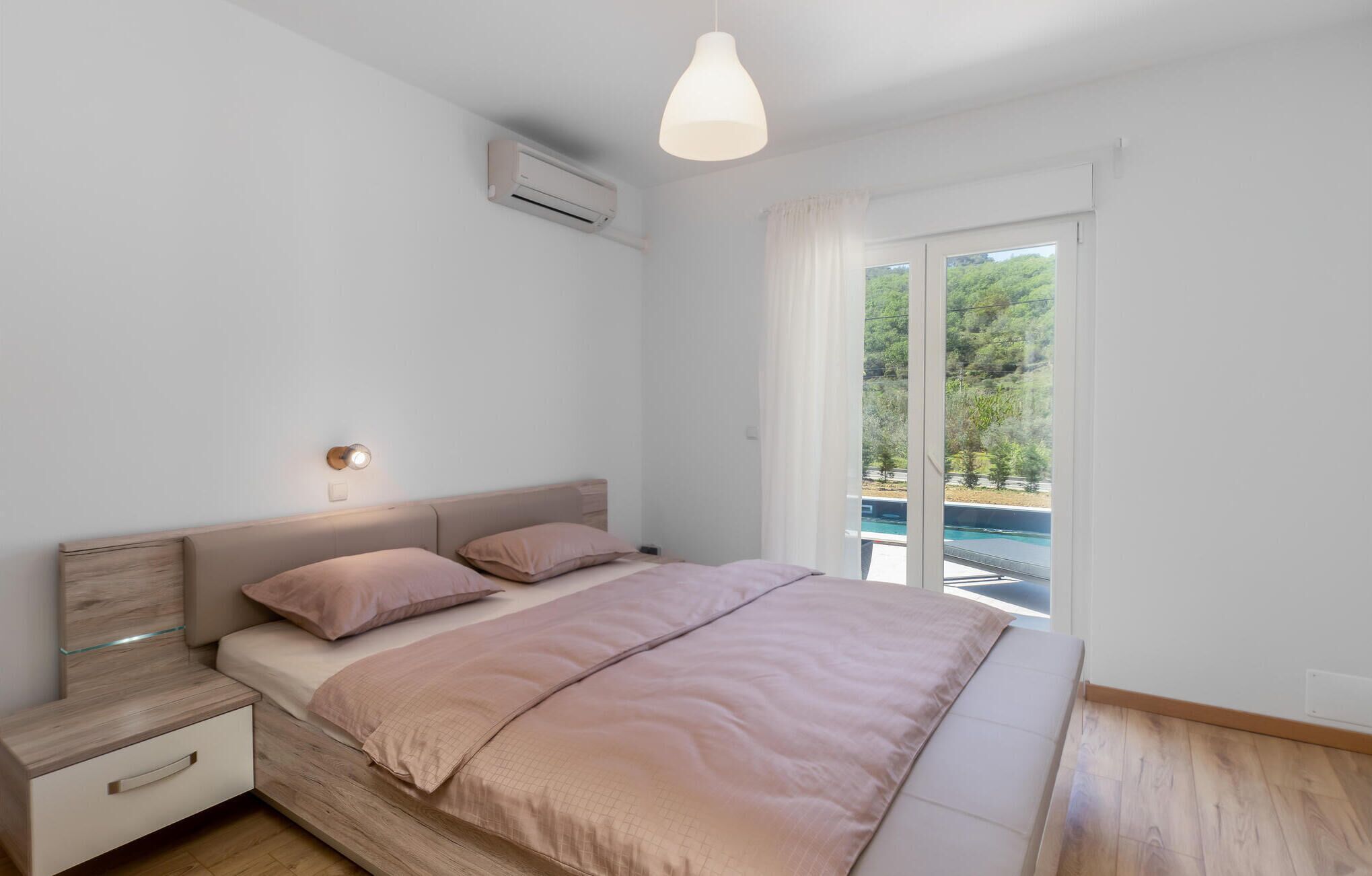 A bedroom with a bed, bedside tables, an air conditioning unit, a pendant light, and a view of a pool through a sliding glass door.