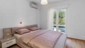 A bedroom with a bed, bedside tables, an air conditioning unit, a pendant light, and a view of a pool through a sliding glass door.