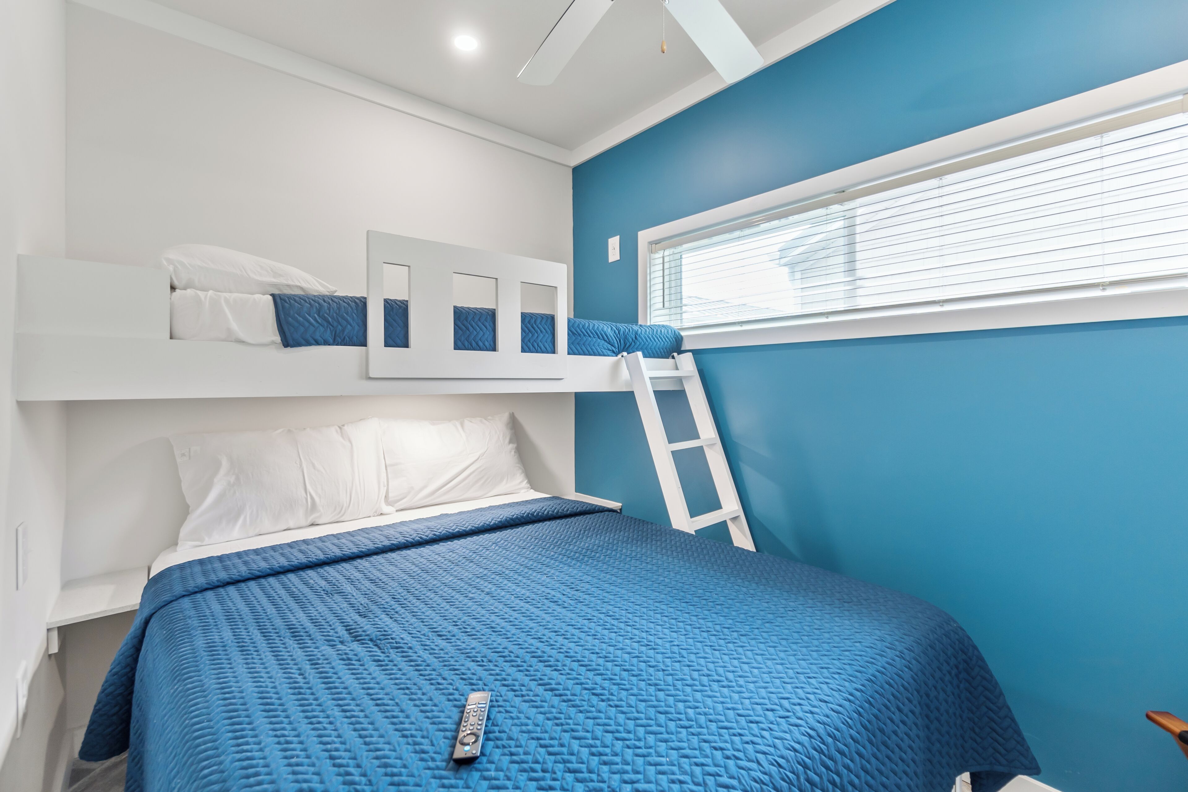A bedroom with a bunk bed, a ladder, a window with blinds, and a blue quilted bedspread.