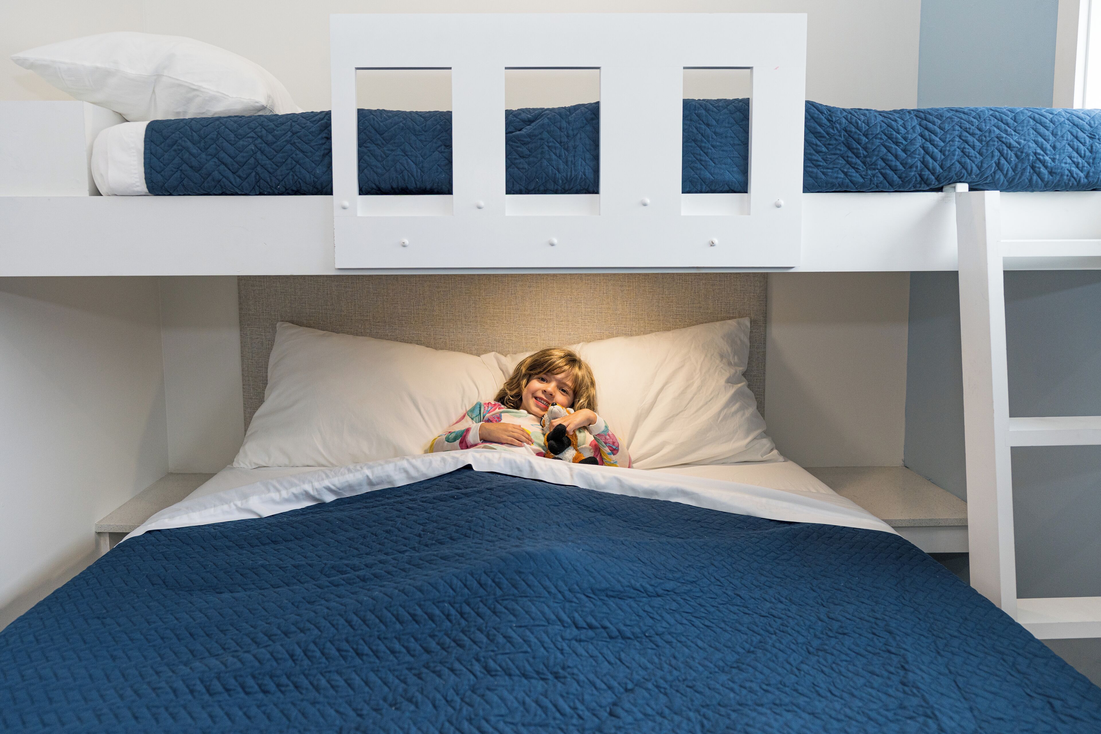 A bunk bed with a white headboard, a lower bed with a blue quilt, and a child reading a book.