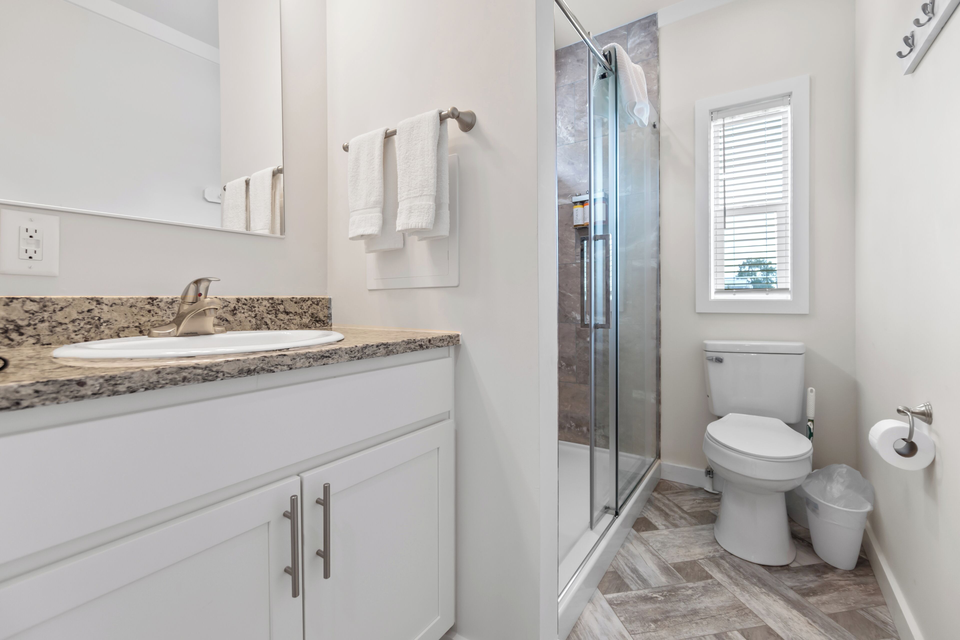 A modern bathroom with a granite countertop sink, white cabinets, a glass shower enclosure, and a toilet.