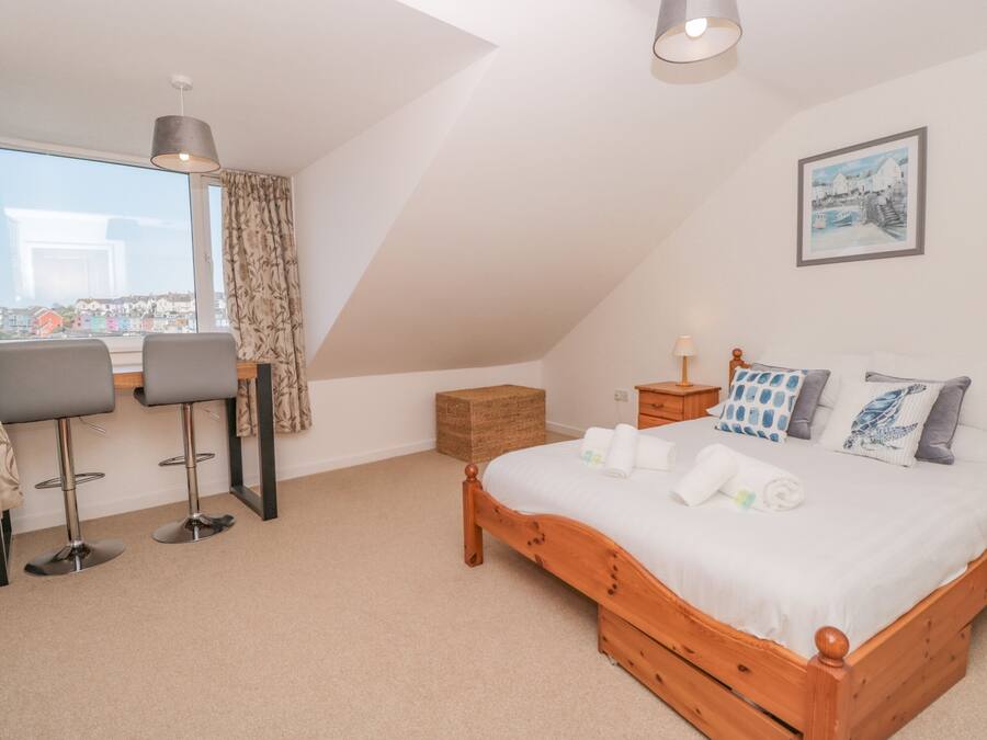 A bedroom with a bed, a desk with two chairs, a window with curtains, a bedside table with a lamp, and a framed picture on the wall.