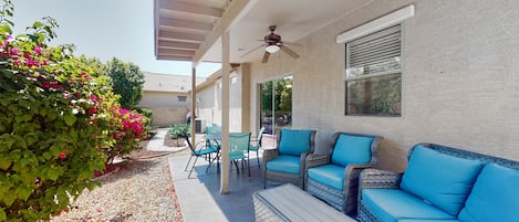 Relax in the shaded patio area with a wicker sofa and armchairs, surrounded by lush greenery and a stucco wall.