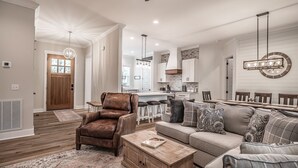 Een ruime woonkamer met een bruine leren fauteuil, een beige bank met opvallende kussens, en een houten salontafel. Achtergrond toont een keuken met witte kastjes en een tegelwand van baksteen.