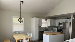 A compact kitchen with white cabinets, a wooden dining table, and a gray sofa. - Peaceful 3-Bedroom Beach Cottage with Deck, Beach Parking Pass & Outdoor Shower (South Kingstown)