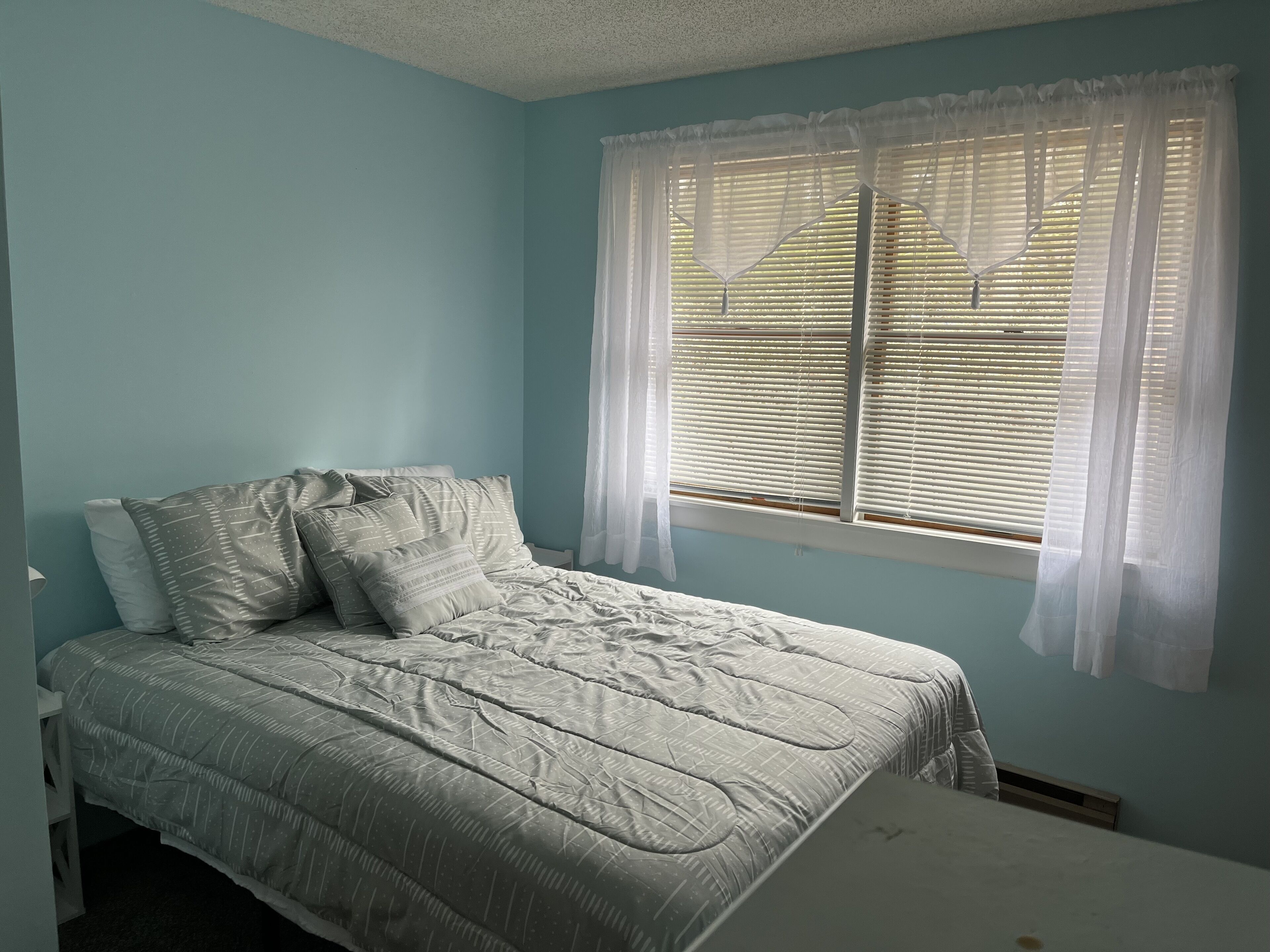A bedroom with a bed, a window with blinds, and a white nightstand.