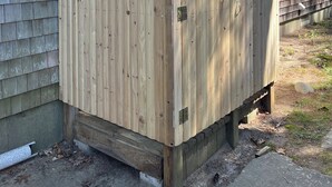A wooden fence with a metal latch, attached to a house with a window and a door. - Peaceful 3-Bedroom Beach Cottage with Deck, Beach Parking Pass & Outdoor Shower (South Kingstown)