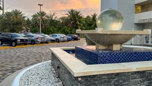Un parking rempli de voitures, un élément décoratif avec de l’eau et un bâtiment doté de balcons.