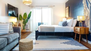 Une chambre moderne avec un lit, des tables de chevet, une télévision, une plante et une grande fenêtre.