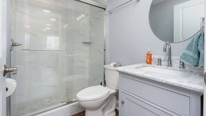 A modern bathroom with a glass-enclosed shower, a white toilet, a marble sink, and a round mirror.
